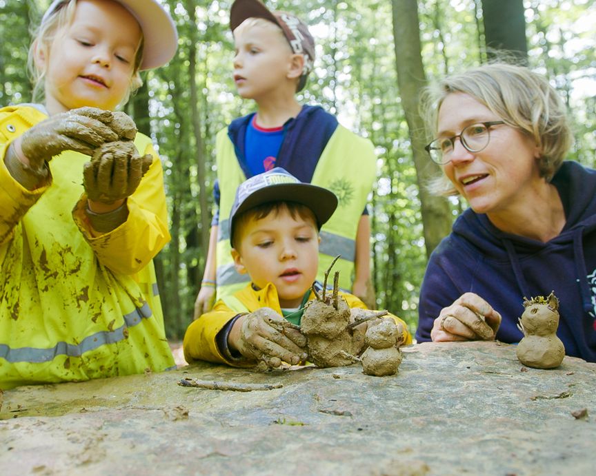 Drei Kinder und eine Erwachsene formen Figuren aus Lehm im Wald
