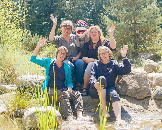 Teamfoto: 4 Frauen sitzen in der Natur und winken.