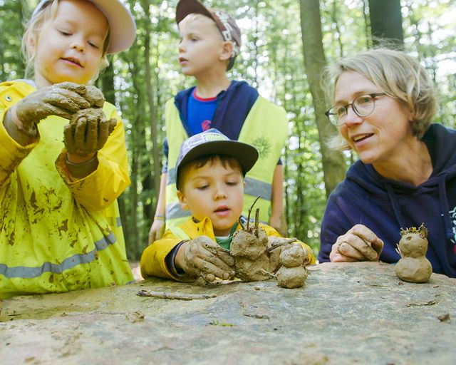 Drei Kinder und eine Erwachsene formen Figuren aus Lehm im Wald